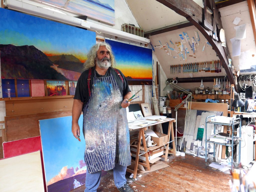 Artist Tom Rickman in his studio, wearing a paint stained apron surrounded by colourful paintings of twilight, he has long white hair & a beard..